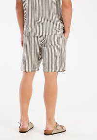 Shorts rayés en coton beige et noir avec une taille élastique et une poche arrière. Le modèle porte des sandales marron à enfiler.