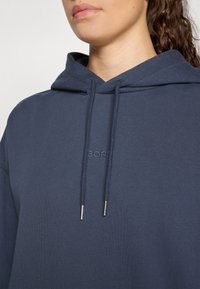 Felpa blu navy realizzata in tessuto morbido con cappuccio regolabile tramite cordini e con aglette argentate; presenta un logo impresso sul fronte.