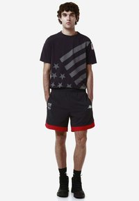 Maglietta nera con design di stelle e strisce grigie, abbinata a pantaloni corti neri con bordo rosso, dettaglio del logo e tasche. Sneakers nere.