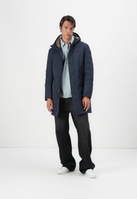 Parka blu navy con zip e chiusura a bottoni a pressione, con fodera interna grigia, tasche frontali e cappuccio. Indossata sopra una camicia abbottonata.