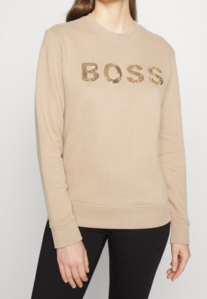 Béžová mikina s dlouhým rukávem a zlatými flitry ve tvaru nápisu "BOSS" nošená osobou v černých kalhotách na jednoduchém pozadí.