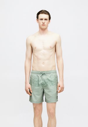 Giovane uomo dalla pelle chiara, capelli castani, in piedi a torso nudo con pantaloncini da bagno verde chiaro a fantasia con coulisse.