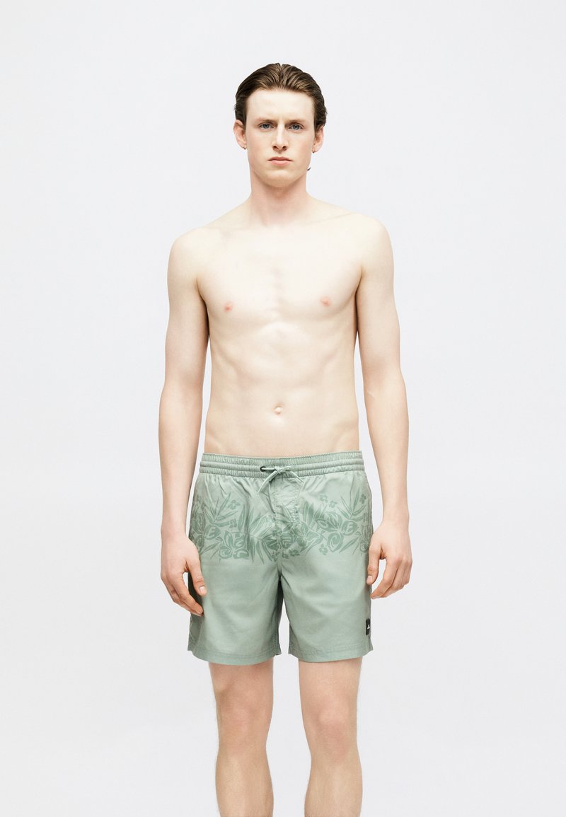 Νεαρός άνδρας με ανοιχτόχρωμο δέρμα και καστανά μαλλιά, όρθιος χωρίς μπλούζα, φορώντας ανοιχτοπράσινα μαγιό με σχέδια και κορδόνι.