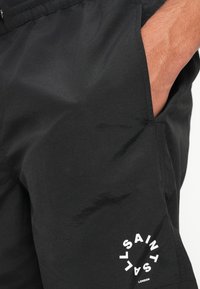 Black shorts made from lightweight fabric, featuring an elastic waistband and side pockets. White circular logo printed on the hem.