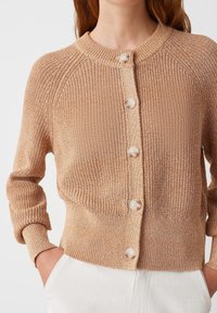 Beige Strickjacke mit rundem Halsausschnitt, Knopfleiste und gerippten Bündchen. Sie verfügt über eine glatte Textur und fünf hellfarbige Knöpfe.