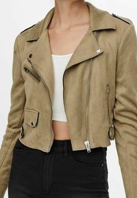 Kvinna klädd i en kortad beige mockabikerjacka med silverdragkedjor, i kombination med en vit ribbad crop top och höga svarta jeans.