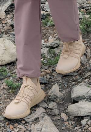 Persona con zapatillas beige y pantalones malva caminando sobre un terreno rocoso e irregular con pequeñas plantas verdes dispersas alrededor.