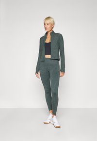 Ensemble de sport vert foncé avec une veste zippée et des leggings ajustés. Tissu lisse et texturé, manches longues avec trous pour les pouces ; accompagné de baskets blanches.