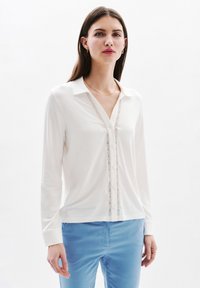 Witte blouse met lange mouwen en een kraag, voorzien van kantdetails aan de voorkant, gecombineerd met blauwe broek. Gladde stofstructuur.