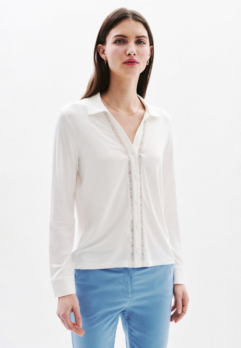Witte blouse met lange mouwen en een kraag, voorzien van kantdetails aan de voorkant, gecombineerd met blauwe broek. Gladde stofstructuur.