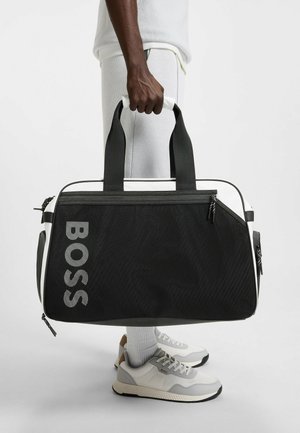 Sac de sport noir et blanc avec un corps texturé noir, des poignées rembourrées et un grand logo argenté "BOSS", disposant de compartiments zippés.