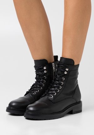 Zwarte leren enkellaarsjes met veter en ritssluiting aan de zijkant, gedragen op blote benen, voorzien van metalen veteroogjes en dikke rubberen zolen.