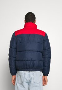 Man i en marinblå och röd pufferjacka samt ljusblå jeans stående med ryggen mot kameran mot en enfärgad bakgrund.