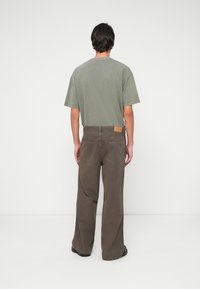 Hombre de pie de espaldas, vistiendo una camiseta verde oliva y pantalones marrones holgados, sobre un fondo blanco liso.