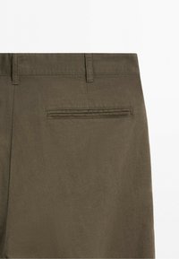 Baksidan av bruna byxor som visar bältesöglor och en enkel pipficka på höger sida mot en vit bakgrund.
