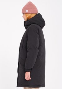 Zwarte pufferjas met een capuchon, voorzien van een voorzak en elastische manchetten. Gecombineerd met een geribbelde roze beanie, schuin gedragen.