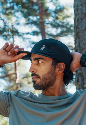 Muž s vousy si venku v lesním prostředí upravuje černou čepici, má na sobě světle modrou košili a černé hodinky na zápěstí.