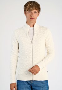 Vit stickad zip-up kofta med diamantmönster, ribbad krage och ärmslut, samt en slät textur, i kombination med blå denimjeans.