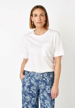 Woman with short brown hair wearing a white short-sleeve shirt with pocket and blue tropical print pants, standing with one hand in pocket.