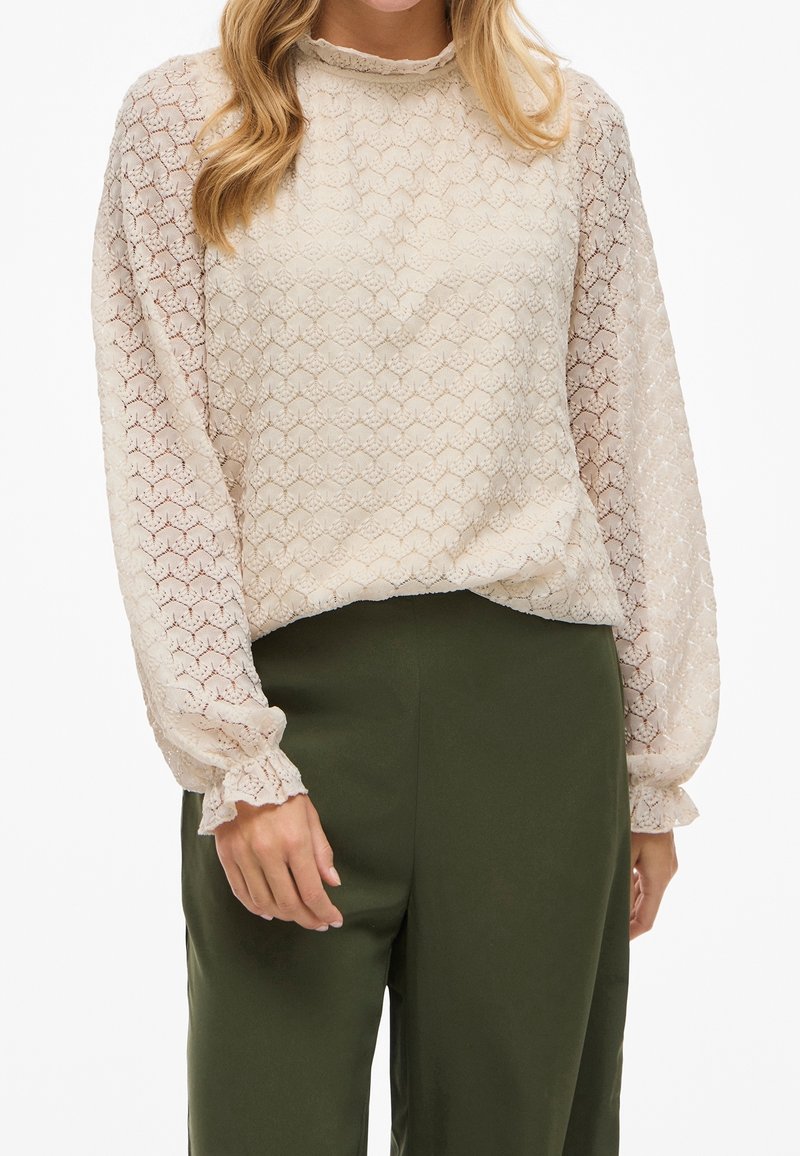 Femme portant un chemisier en dentelle crème transparente à manches longues avec des poignets à volants, associé à un pantalon taille haute vert olive.