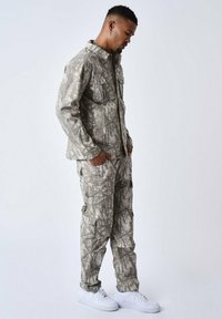 Camouflage-jack en -broek in lichtgrijze tinten, gemaakt van stof met een gladde textuur. Gecombineerd met witte sneakers.