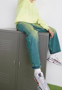 Sudadera con capucha de color amarillo neón, jeans azules desgastados y zapatillas blancas con acentos rojos y negros, sentados en un casillero gris.