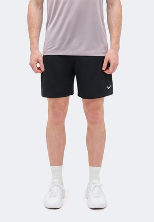 Homme portant un short noir Nike, une chemise de couleur claire, des chaussettes blanches et des chaussures de sport blanches, se tenant devant un fond clair uni.
