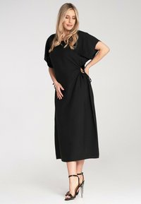 Schwarzes Midi-Kleid mit kurzen senkrechten Ärmeln, seitlichen Schnürelementen, lockerer Passform und leichtem Material, kombiniert mit hohen Sandalen.