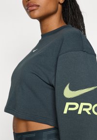 Mørkegrøn cropped sweatshirt lavet af blødt stof, med et hvidt Nike-logo på brystet og gule "PRO"-tekster på ærmet.