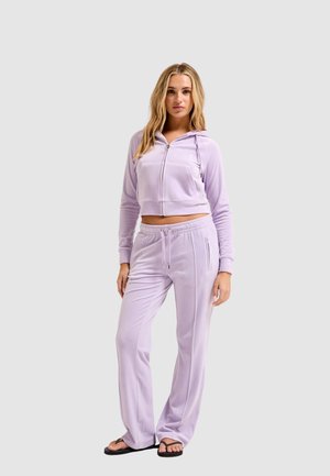 Vrouw staand in lichtpaarse velours zip-up hoodie en bijpassende wijde broek met zwarte slippers op een grijze achtergrond.