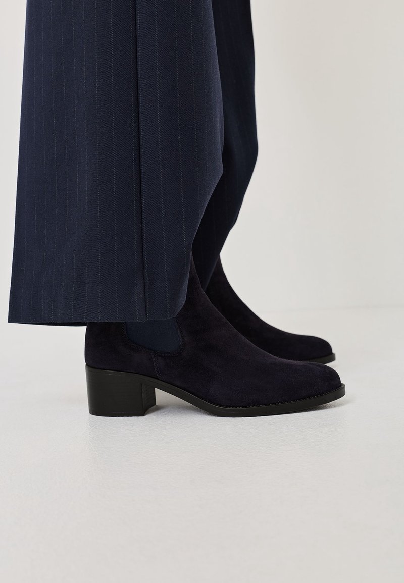 Bottines en daim bleu marine à talons blocs portées avec un pantalon large à fines rayures foncées, vues de profil sur un sol clair.