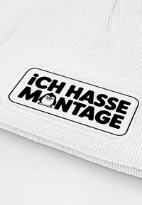White knitted beanie featuring a rectangular patch with the text "Ich hasse Montage" and a cartoon penguin graphic.