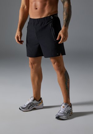 Shorts de sport noirs avec une taille à cordon de serrage et une poche latérale zippée. Portés avec des chaussures de course grises et blanches, avec des accents colorés.