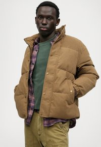 Bruine ribfluwelen pufferjas met drukknoopjes, gedragen over een groene shirt en een geruite flanel, met een relaxte pasvorm en geribbelde manchetten.