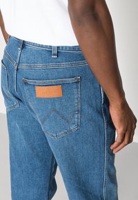 Niebieskie dżinsy z teksturowanym wykończeniem, z skórzaną łatą z logo "Wrangler" i kontrastowymi szwami na tylnych kieszeniach.