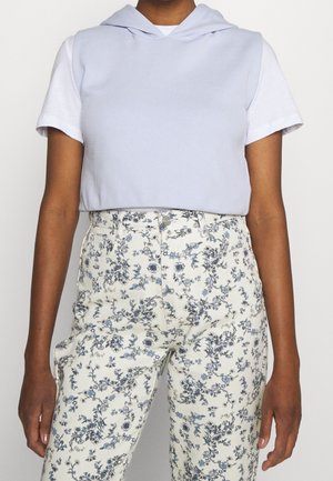 Personne portant une chemise blanche à manches courtes sous un haut sans manches à capuche bleu clair avec un pantalon blanc taille haute à imprimé floral bleu.