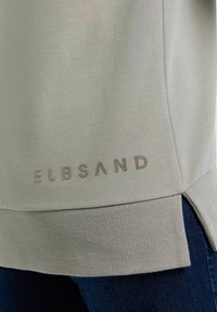 Ourlet de sweat-shirt gris clair avec bord côtelé, fente sur le côté et texte embossé "ELBSAND", associé à un jean bleu foncé.