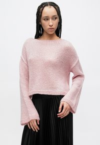 Ανοιχτό ροζ πλεκτό πουλόβερ με κοντό σχέδιο, φαρδιά μανίκια και άνετη γραμμή, σε συνδυασμό με μαύρη πλισέ φούστα.