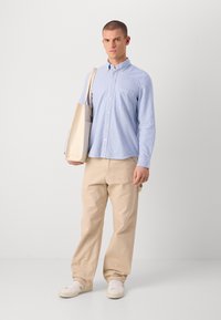 Blå og hvit stripet skjorte med knapper, beige bukser, hvite sneakers, holder en lys farget totebag. Enkel, uformell stil med en avslappet silhuett.