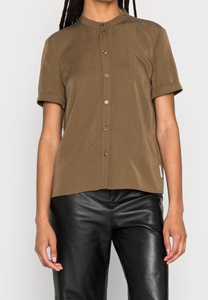 Woman wearing brown short-sleeve button-up shirt and black leather pants, standing with arms relaxed by sides.