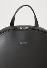 Fekete műbőr hátizsák kerekített formával, cipzáros záródással és ezüst színű fémrészekkel. A Calvin Klein logó a elülső részen domborítva.