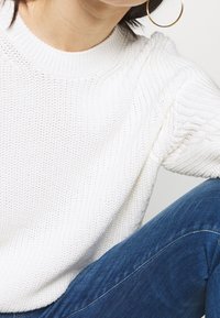 Joven con suéter de punto blanco, jeans azules y grandes pendientes de aro dorados, sentado con el brazo apoyado en la rodilla doblada.