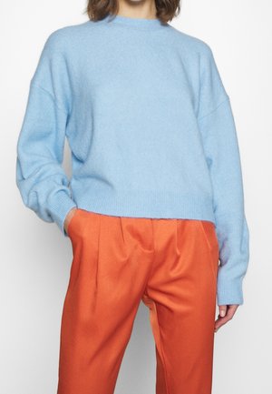 Osoba ubrana w jasnoniebieski puszysty sweter i luźne, jaskrawo pomarańczowe spodnie, z rękami w kieszeniach.