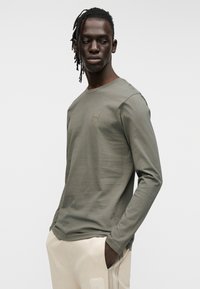 Μακρυμάνικη μπλούζα σε ελαιί πράσινο, στρογγυλή λαιμόκοψη, εφαρμοστή γραμμή, με μικρό λογότυπο στο στήθος. Απαλή υφή από βαμβάκι.