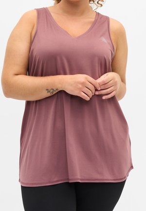 Femme portant un haut mauve sans manches à col en V avec un tatouage au poignet, les mains jointes, et un legging noir sur un fond clair uni.