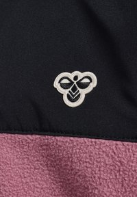 Zwarte stof met een roze fleece paneel met textuur aan de onderkant, voorzien van een wit geborduurd logo dat lijkt op een bij met decoratieve elementen.