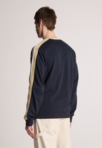 Sweatshirt marine à manches longues, avec des rayures dorées et blanches sur les côtés. Tissu doux, col rond et poignets et ourlet côtelés.