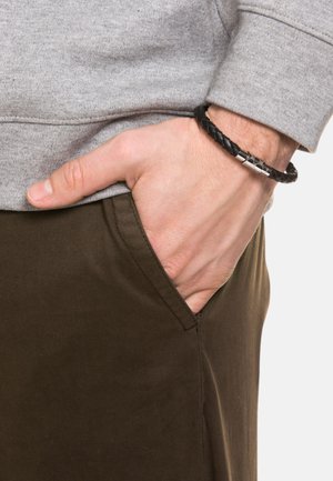 Sudadera gris con cuello redondo, combinada con pantalones marrones oscuros. Pulsera trenzada negra llevada en la muñeca. Mano reposando en el bolsillo.