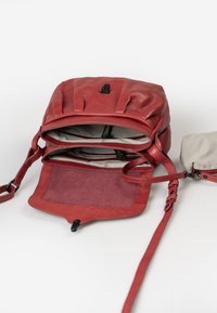 Roter Leder-Rucksack mit gefaltetem Design, ausgestattet mit einer glatten Oberfläche und einem gestreiften Stoffinnenraum. Enthält einen verstellbaren Riemen und eine Reißverschlusstasche.
