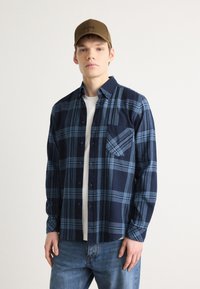 Camisa xy com padrão xadrez em azul-marinho e azul claro, com botões à frente, gola e bolso no peito, usada sobre uma camisete branca, combinada com jeans azuis e um boné castanho.
